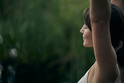 Eine Frau hat ihre Arme nach oben gerichtet und streckt sich, im Hintergrund eine grüne Wiese. Sie hat die Augen geschlossen und entspannt.