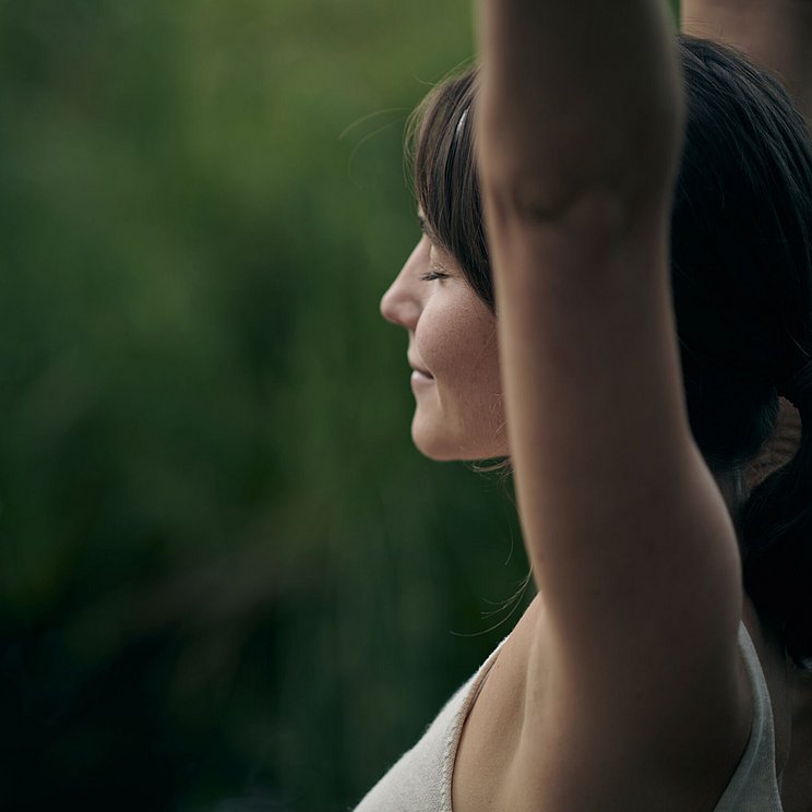 Eine Frau hat ihre Arme nach oben gerichtet und streckt sich, im Hintergrund eine grüne Wiese. Sie hat die Augen geschlossen und entspannt.