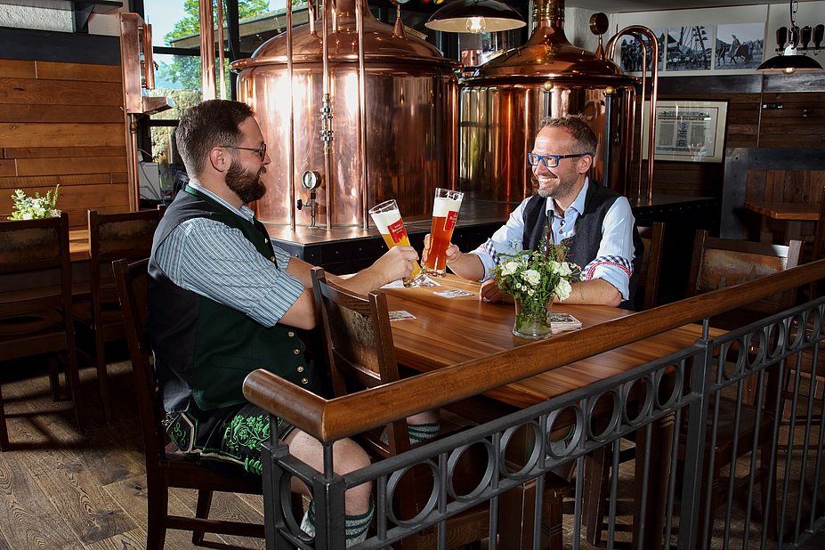 Zwei Männer sitzen in einem Brauhaus und stoßen mit Bier an, sie tragen Tracht