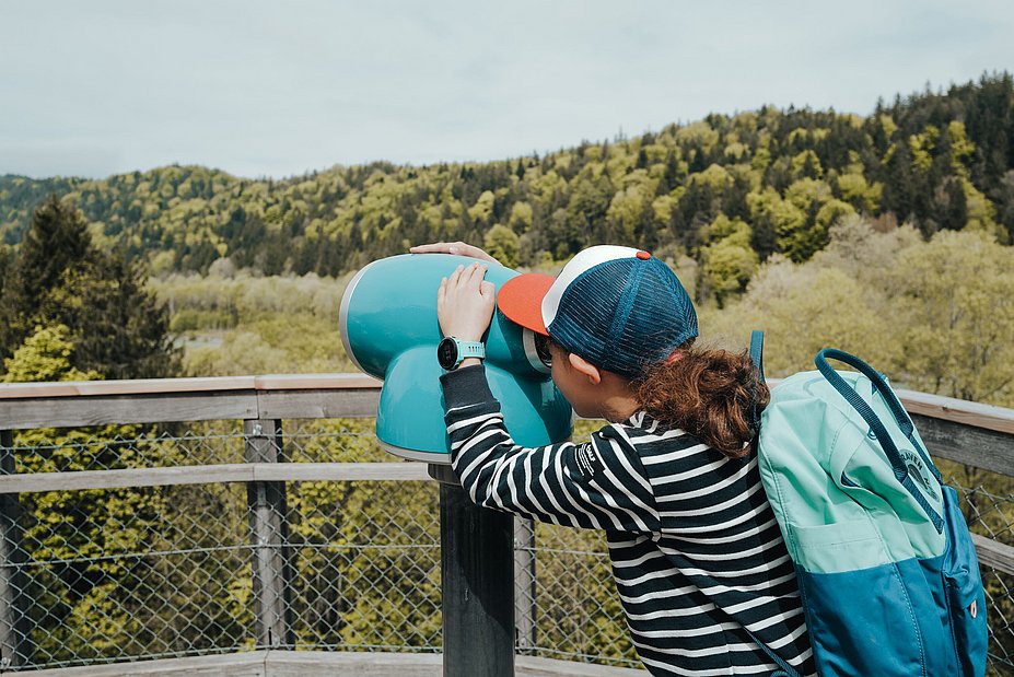 Mädchen mit Käppi schaut durch ein Fernglas über bewaldete Hügel