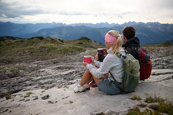 Eine Frau und ein Mann sitzen mit Bechern in der Hand auf dem Gottesackerplateau und sehen in die Berge