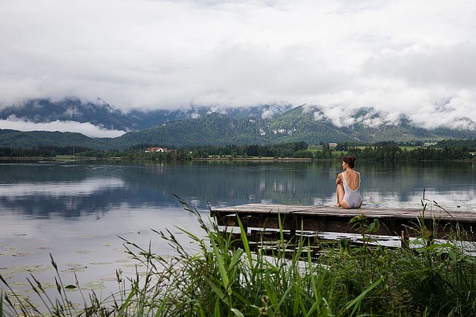 Frau im Badeanzug sitzt auf einem Holzsteg am Hopfensee und blickt übers Wasser, die Berge sind wolkenverhangen
