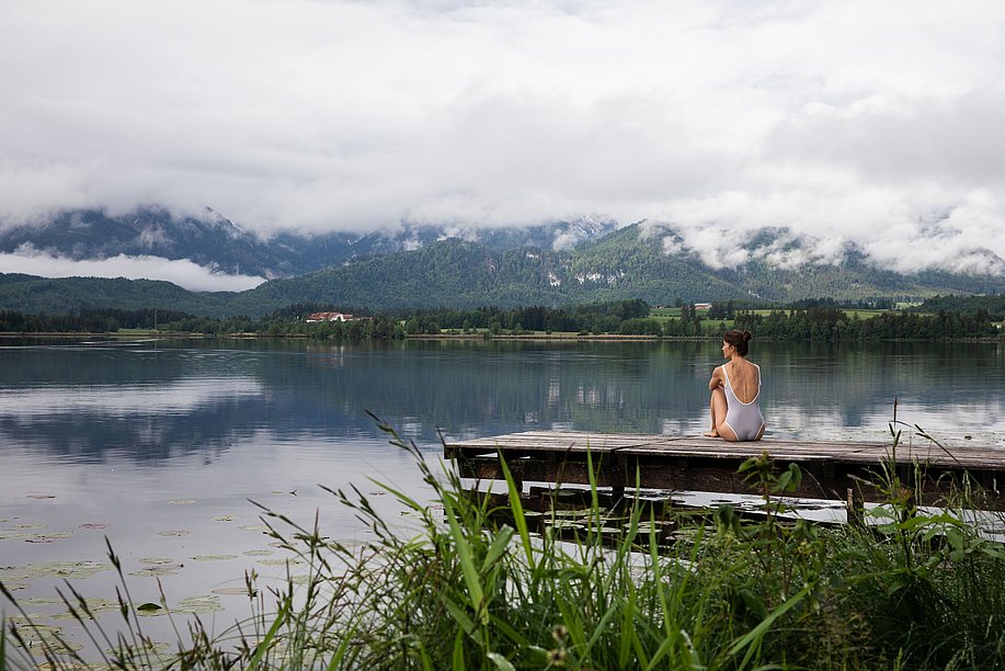 Frau im Badeanzug sitzt auf einem Holzsteg am Hopfensee und blickt übers Wasser, die Berge sind wolkenverhangen
