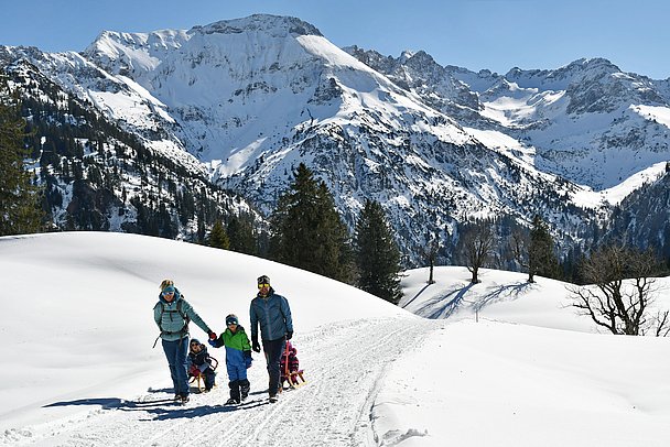 Eltern ziehen zwei Rodel eine präparierte Strecke hinauf, ein Kind geht an der Hand, im Hintergrund verschneites Bergpanorama