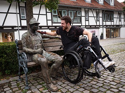 Moritz Brückner im Rollstuhl legt die Hand auf die Statue eines Mannes, der auf einer Holzbank sitzt