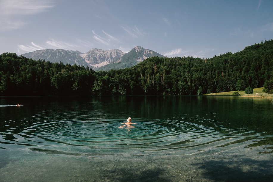 Frau schwimmt im Alatsee mit bewaldetem Ufer und Bergen im Hintergrund