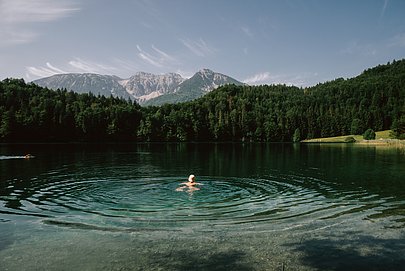 Frau schwimmt im Alatsee mit bewaldetem Ufer und Bergen im Hintergrund