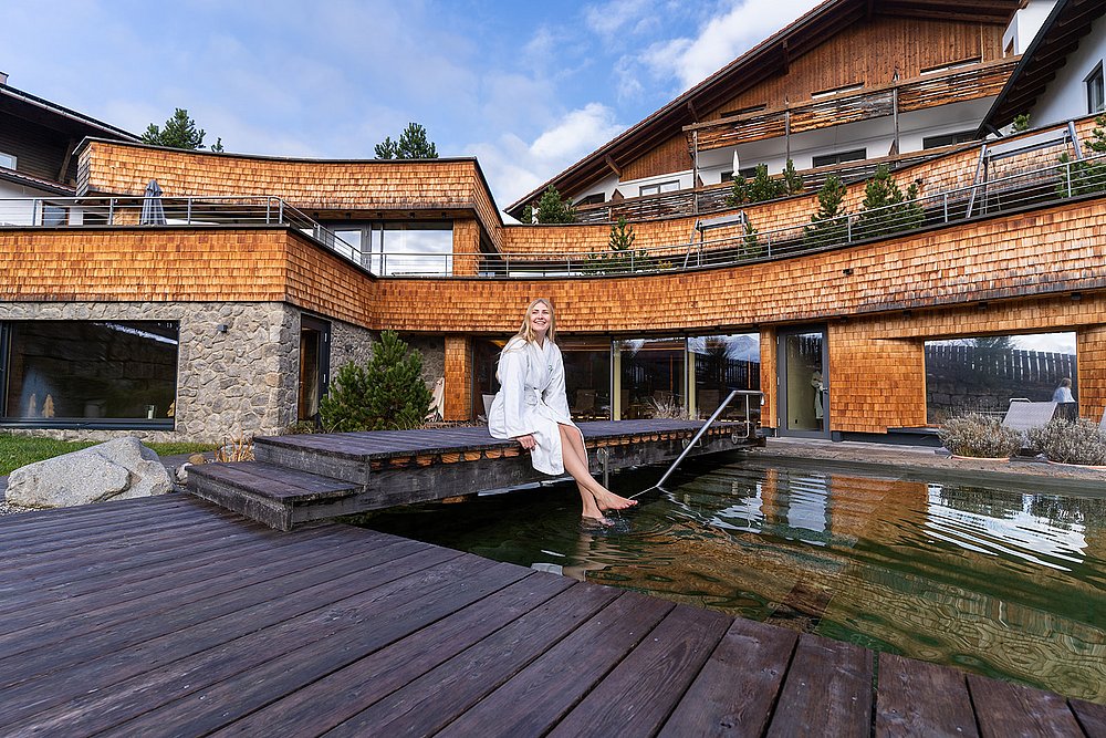 Frau im Bademantel auf einem Holzsteg sitzend mit den Füßen im Pool vor dem Biohotel Eggensberger