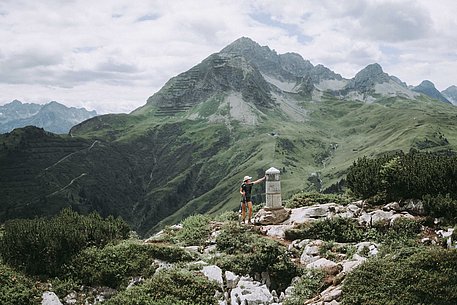 Wanderin lehnt am Grenzstein, der am südlichsten Punkt Deutschlands steht, im Hintergrund Berge