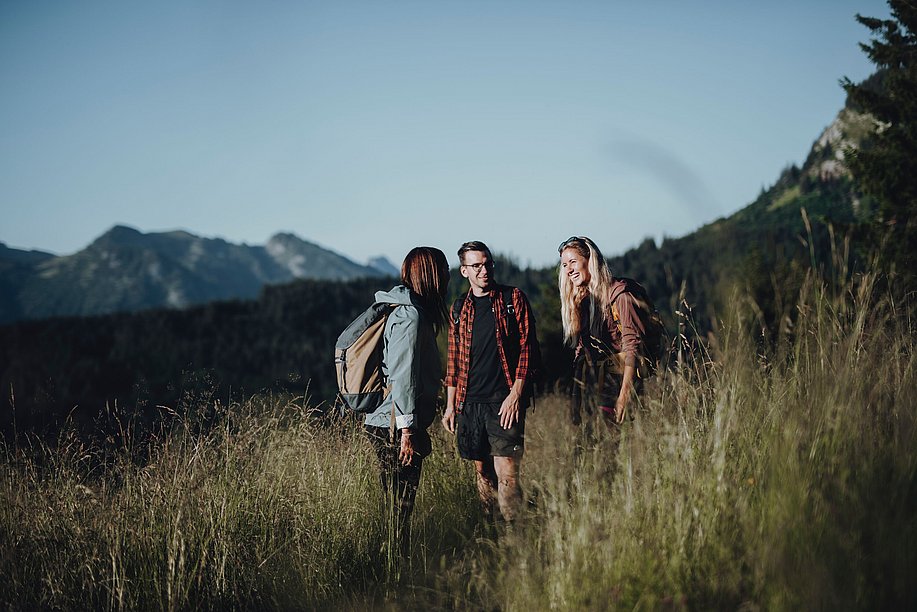 Zwei junge Frauen und ein Mann auf einer Bergwiese unterhalten sich und lachen