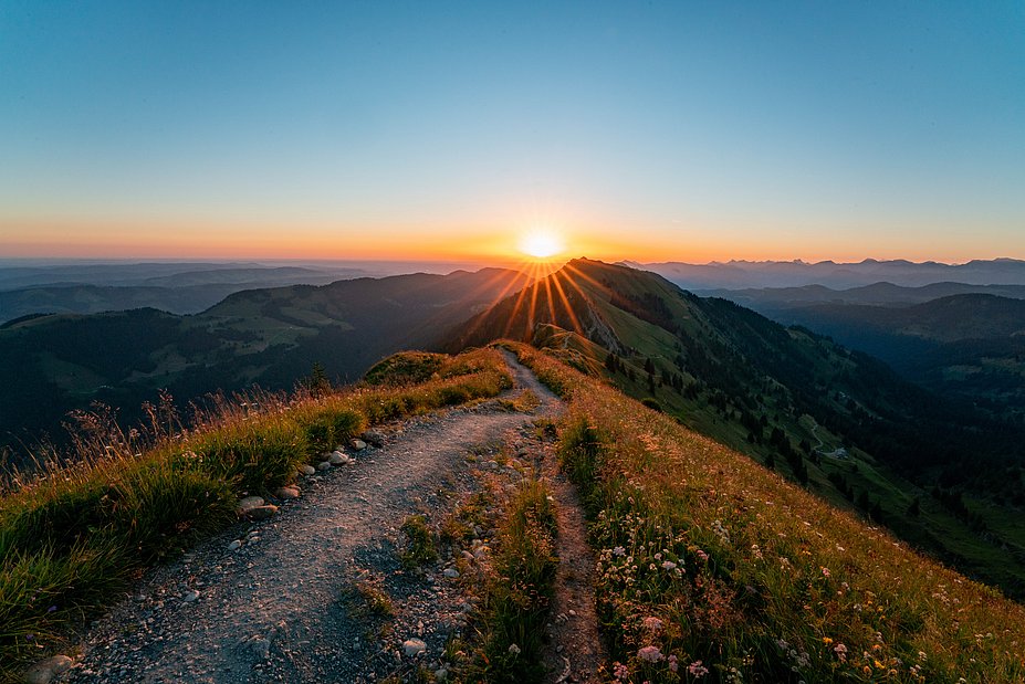 Ein Kammweg zwischen Wiesen, im Hintergrund geht die Sonne über den Bergen auf