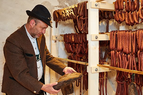 Traditionell gekleideter Mann vor einem Gestell mit trocknenden Würsten