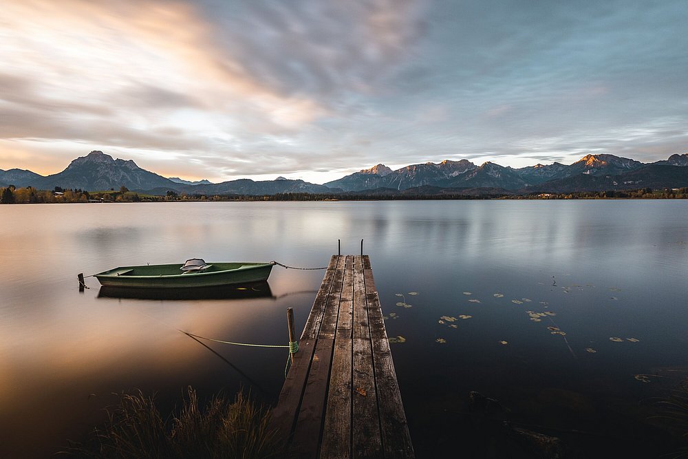 Holzsteg am Hopfensee mit einem grünem Fischerboot, die Ammergauer Alpen im Hintergrund im Licht der untergehenden Sonne