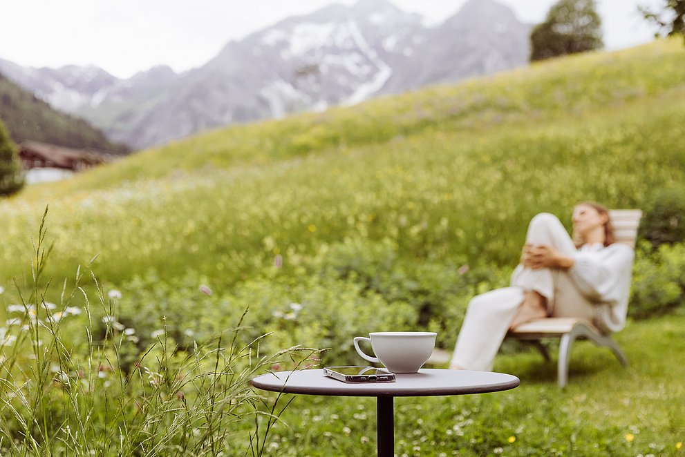 Frau im Liegestuhl vor Bergpanorama, im Vordergrund ein kleiner Tisch mit Teetasse und Handy