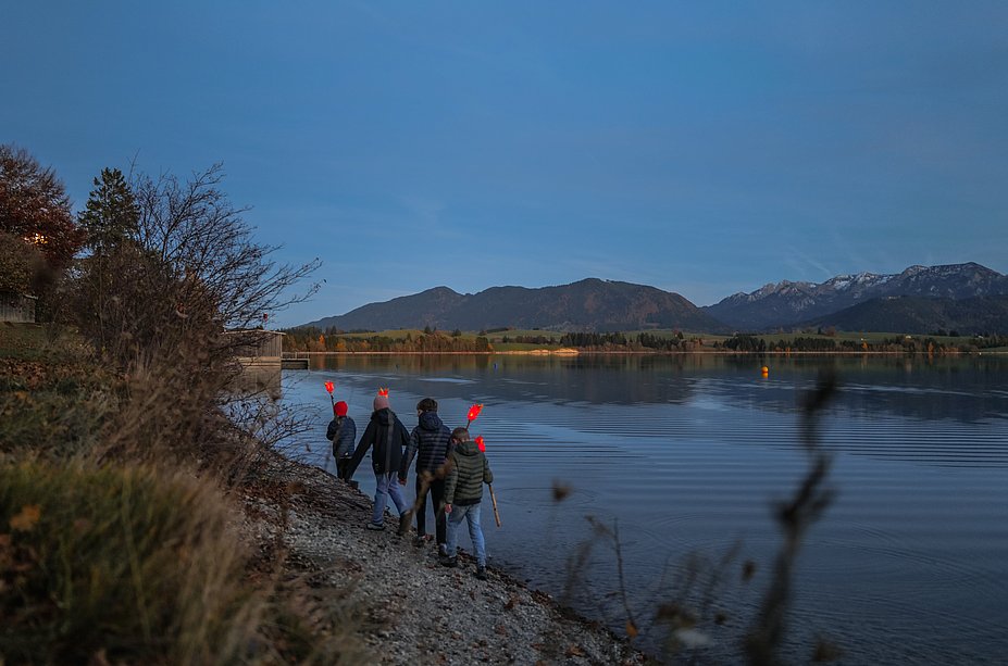 Vier Kinder wandern in der Dämmerung mit selbstgebastelten leuchtenden Fackeln an einem Seeufer entlang