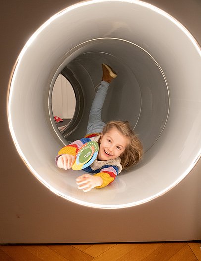 Kind mit Rätselscheibe in der Hand in einem Loch in der Wand, durch das man krabbeln kann, im Stadtmuseum Isny