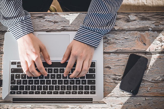 Laptop von oben auf einem Holztisch, Hände auf der Tastatur, daneben eine Handy