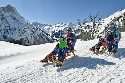 Eltern mit drei Kindern beim Rodeln vor verschneiter Bergkulisse