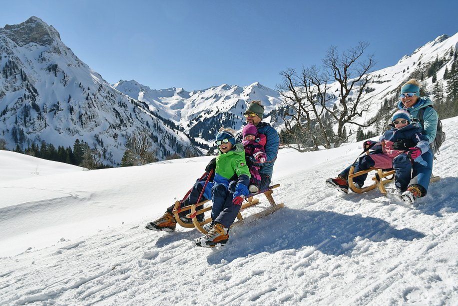 Eltern mit drei Kindern beim Rodeln vor verschneiter Bergkulisse