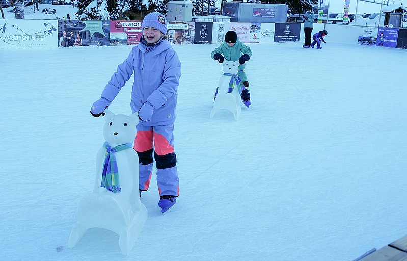Zwei Kinder toben sich auf der Eisbahn im Tannheimer Tal aus. Sie haben einen Eisbär als Lernhilfe.