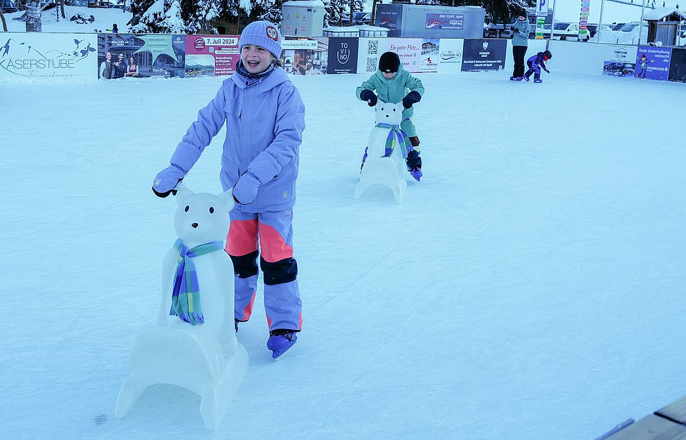 Zwei Kinder toben sich auf der Eisbahn im Tannheimer Tal aus. Sie haben einen Eisbär als Lernhilfe.