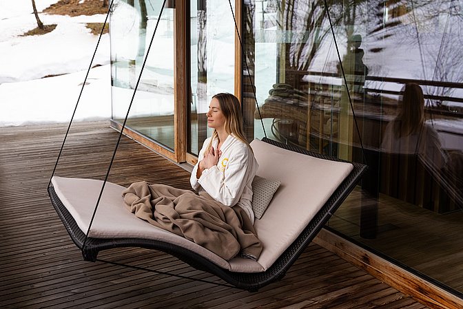 Frau im Bademantel sitzt im Hängesitz auf einer Terrasse vor verschneiter Landschaft