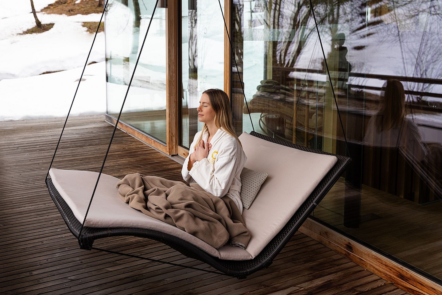 Frau im Bademantel sitzt im Hängesitz auf einer Terrasse vor verschneiter Landschaft