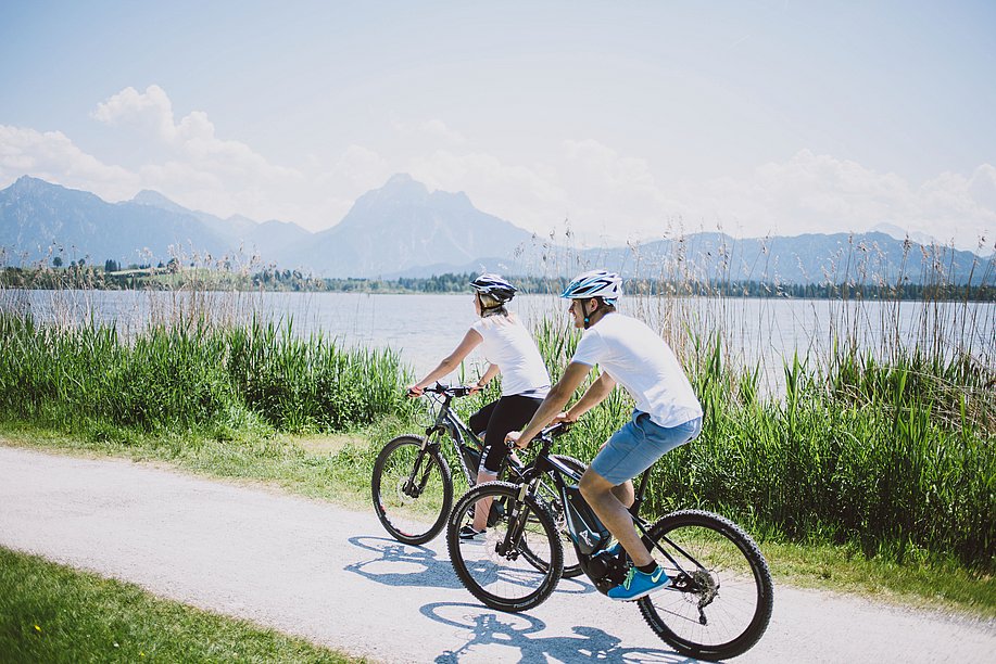 Mann und Frau auf Fahrrädern auf einem Weg entlang eines Sees, im Hintergrund die Berge
