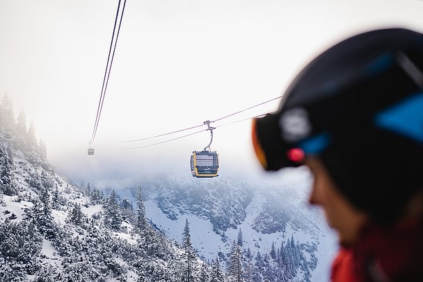 Kopf eines Skifahrers im Vordergrund, im Hintergrund die Gondeln einer Bergbahn