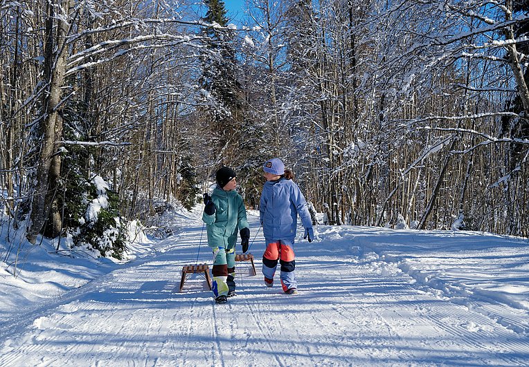Zwei Kinder ziehen einen Holzschlitten durch die Winterlandschaft. 