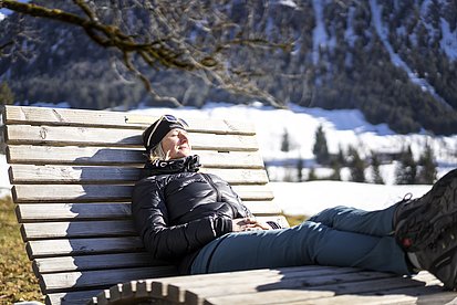 Frau liegt entspannt auf einer Holzbank in der Sonne, dahinter Winterlandschaft