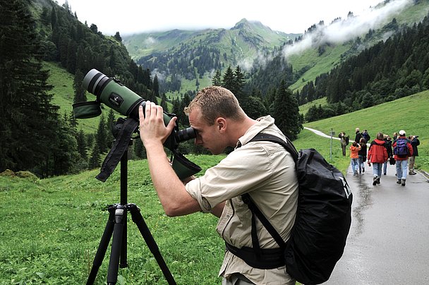 Ein Mann guckt durch ein großes Fernrohr nach oben, im Hintergrund wandert eine Gruppe durchs Tal