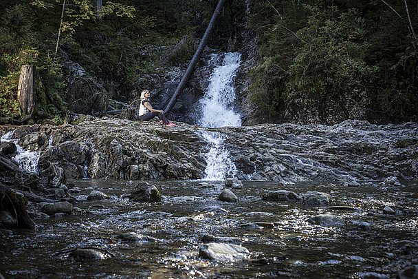 Frau sitzt auf einem Felsen vor einem Wasserfall im Wald