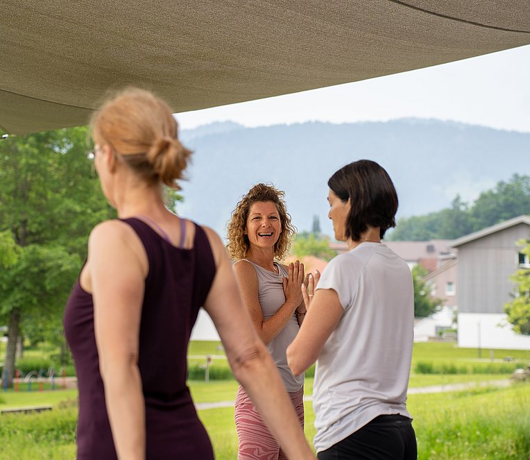 Im Vordergrund zwei Frauen von hinten, dahinter lacht eine Frau in die Kamera, die Hände vor der Brust aufeinander gelegt