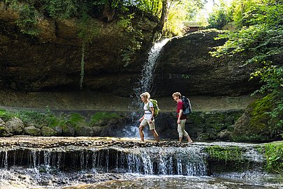 Zwei Wanderer gehen vor einem Wasserfall durchs Wasser