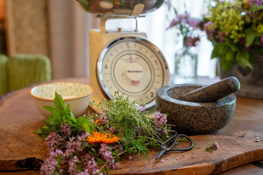 Waage, Mörser und Schale, davor ein Holzbrett mit mit Kräutern und Blüten