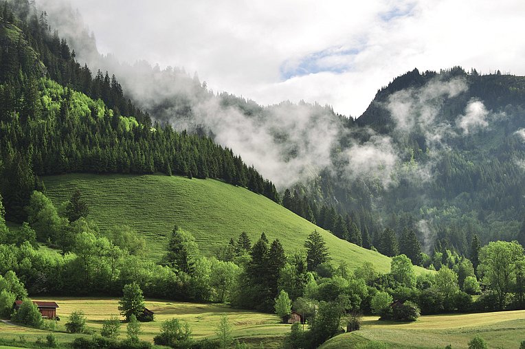 Wolken zwischen Bergen mit Wiesen, Wäldern und kleinen Holzhütten