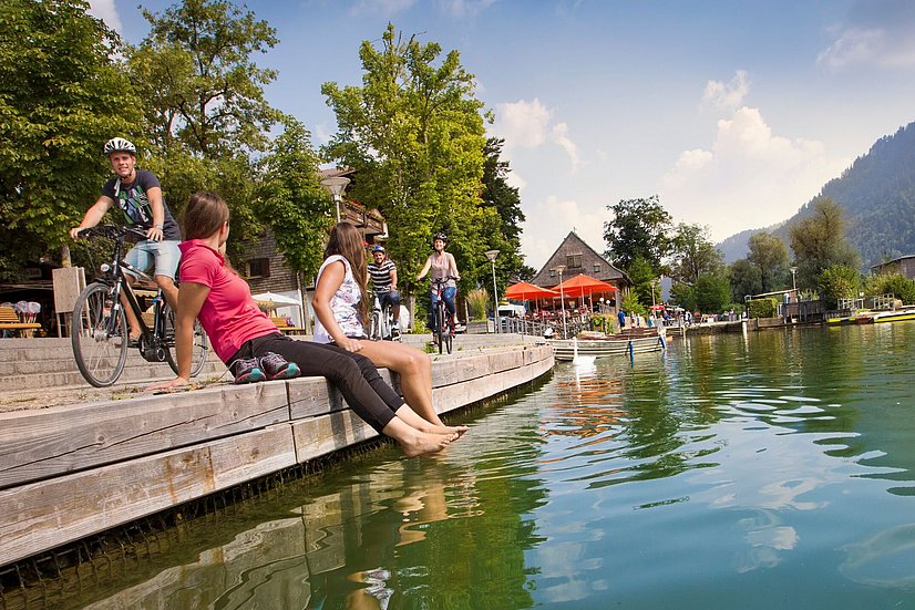 Zwei Frauen sitzen am Ufer des Alpsees mit den Füßen im Wasser, dahinter drei Fahrradfahrer
