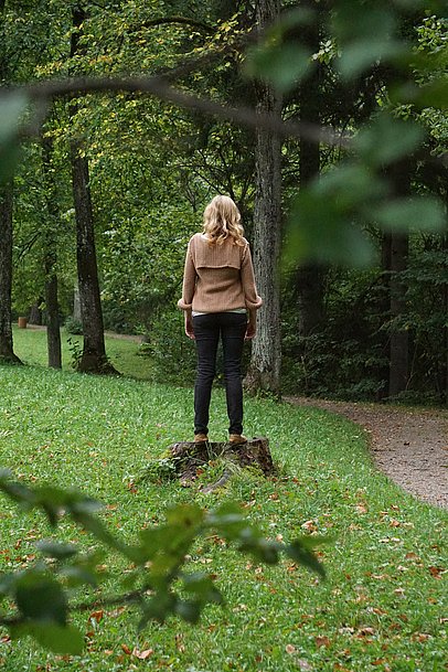Eine Frau von hinten auf einem Baumstumpf zwischen Wiesen und Bäumen