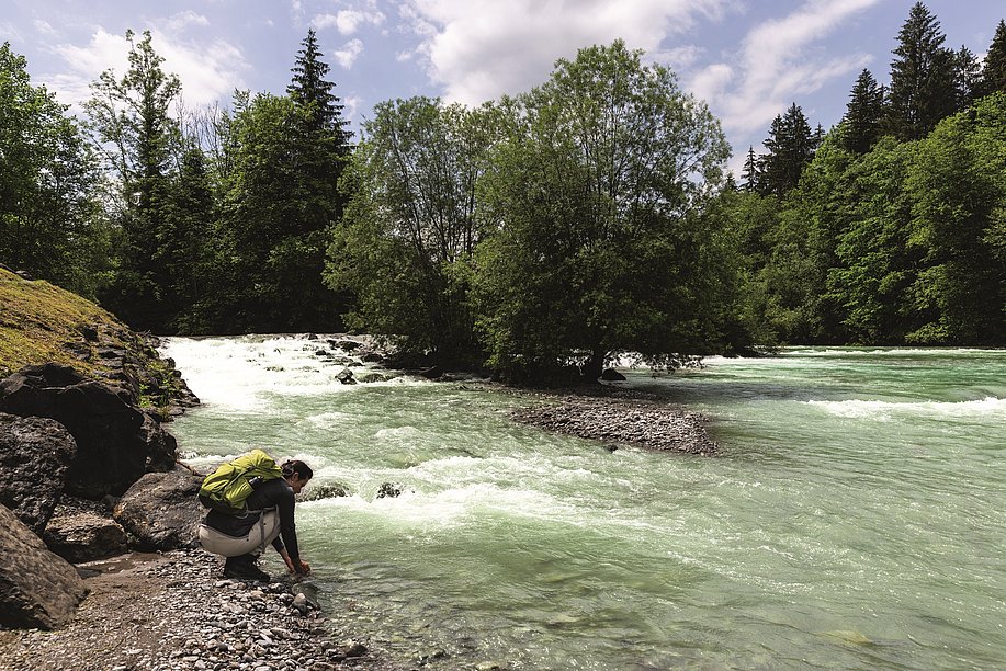 Eine Frau mit Rucksack hält ihre Hände in das Wasser eines Flusses, an den Ufern Felsen und Bäume