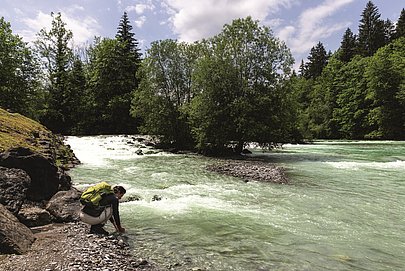 Eine Frau mit Rucksack hält ihre Hände in das Wasser eines Flusses, an den Ufern Felsen und Bäume