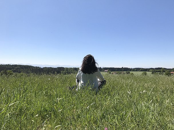 Frau sitzt in einer Wiese, im Hintergrund die Alpen