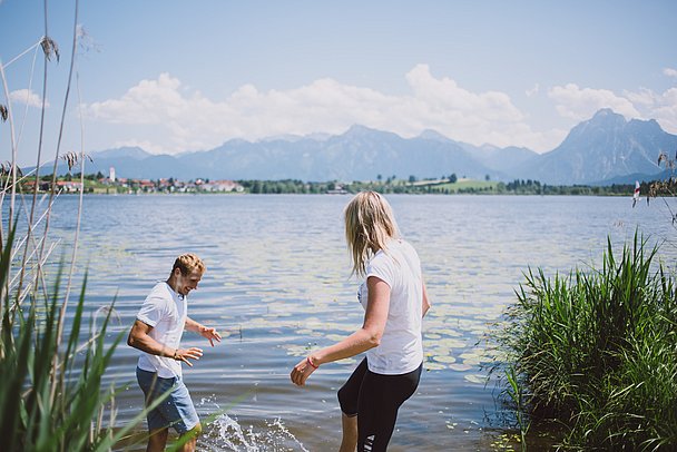 Mann und Frau mit den Füssen im Wasser eines Sees, im Hintergrund die Berge