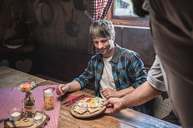 In der Stube einer Hütte bekommt ein Mann eine Brotzeit serviert