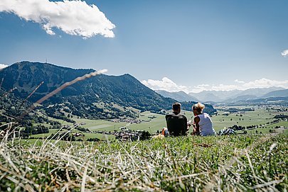 Paar sitzt auf einer Wiese auf der Gebhardshöhe, im Hintergrund die Allgäuer Alpen