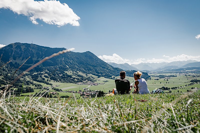 Paar sitzt auf einer Wiese auf der Gebhardshöhe, im Hintergrund die Allgäuer Alpen