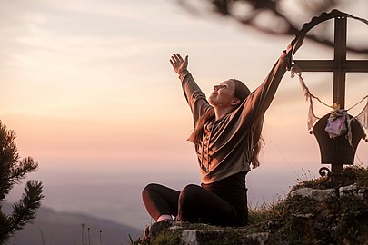 Frau sitzt an einem Gipfelkreuz mit ausgebreiteten Armen Richtung Himmel Frau sitzt an einem Gipfelkreuz mit ausgebreiteten Armen Richtung Himmel