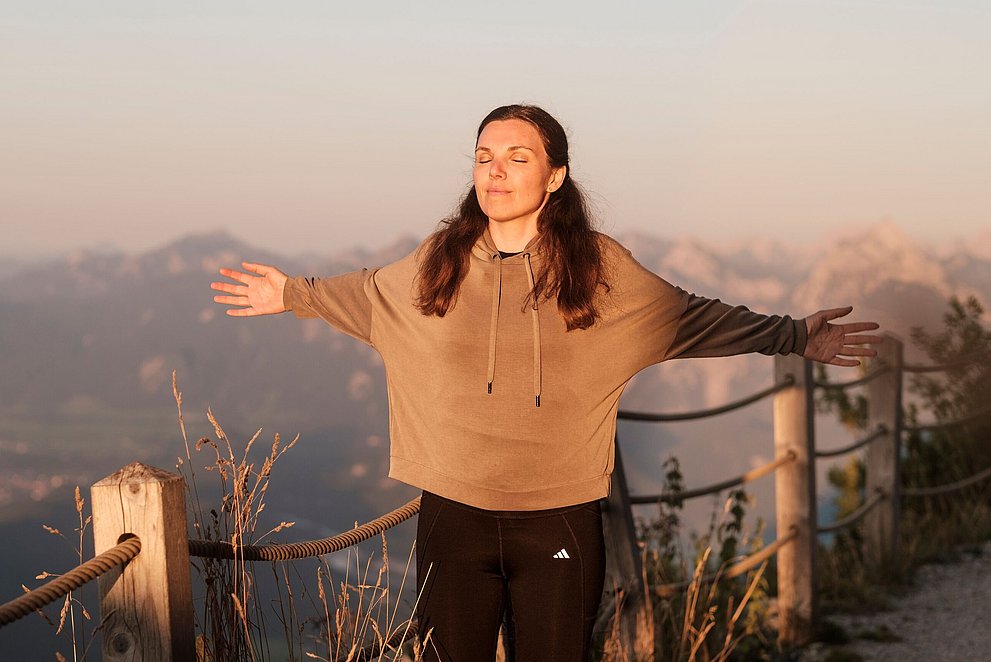 Frau mit zufriedenem Gesicht breitet die Arme zur Seite aus vor einer Berglandschaft