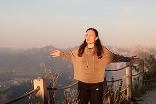 Frau mit zufriedenem Gesicht breitet die Arme zur Seite aus vor einer Berglandschaft