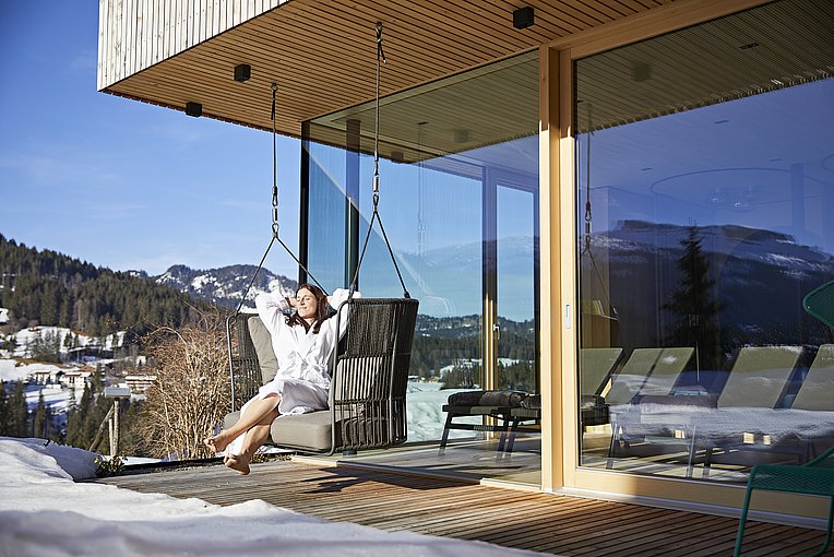 Frau im Bademantel sitzt entspannt in der Sonne auf der Holzterrasse des Biohotels Oswalda Hus, ringsherum Schnee